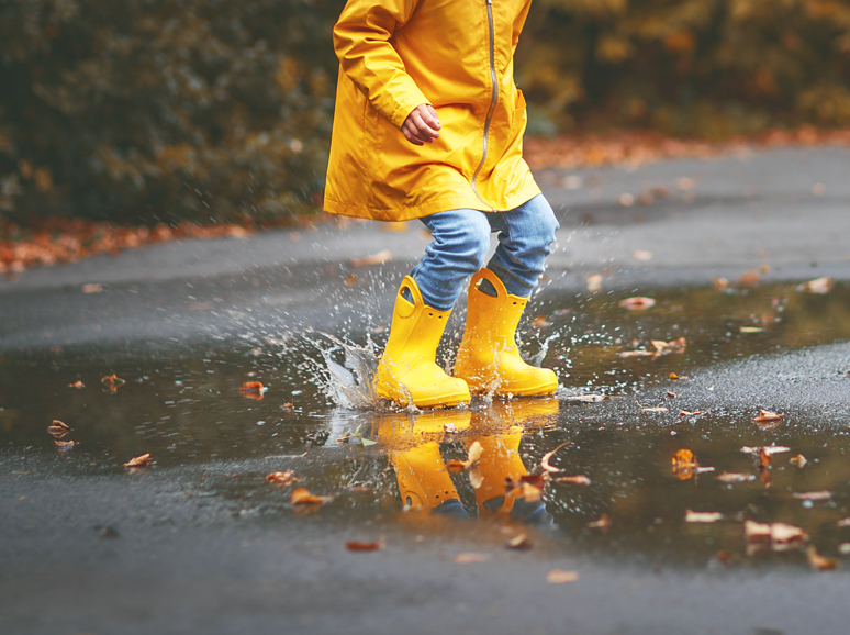 A child in a yellow raincoat and boots is splashing in a puddle